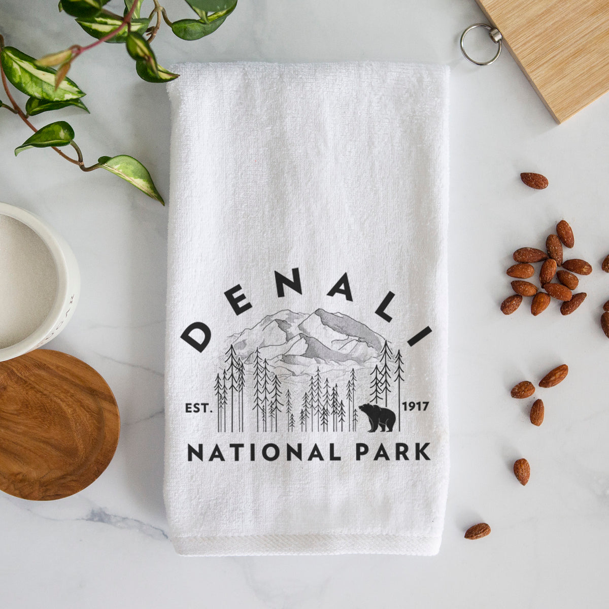 White hand towel printed with a round badge of mountains, pine trees and a bear, with the words Denali National Park, Est. 1917.