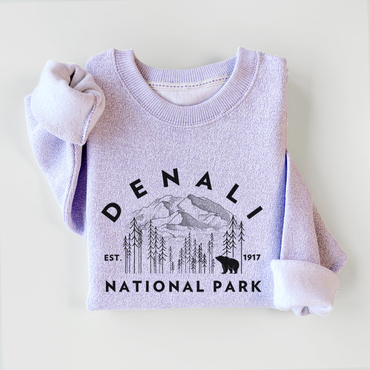Ice purple knit sweatshirt printed with a round badge of mountains, pine trees and a bear, with the words Denali National Park, Est. 1917, styled flat.