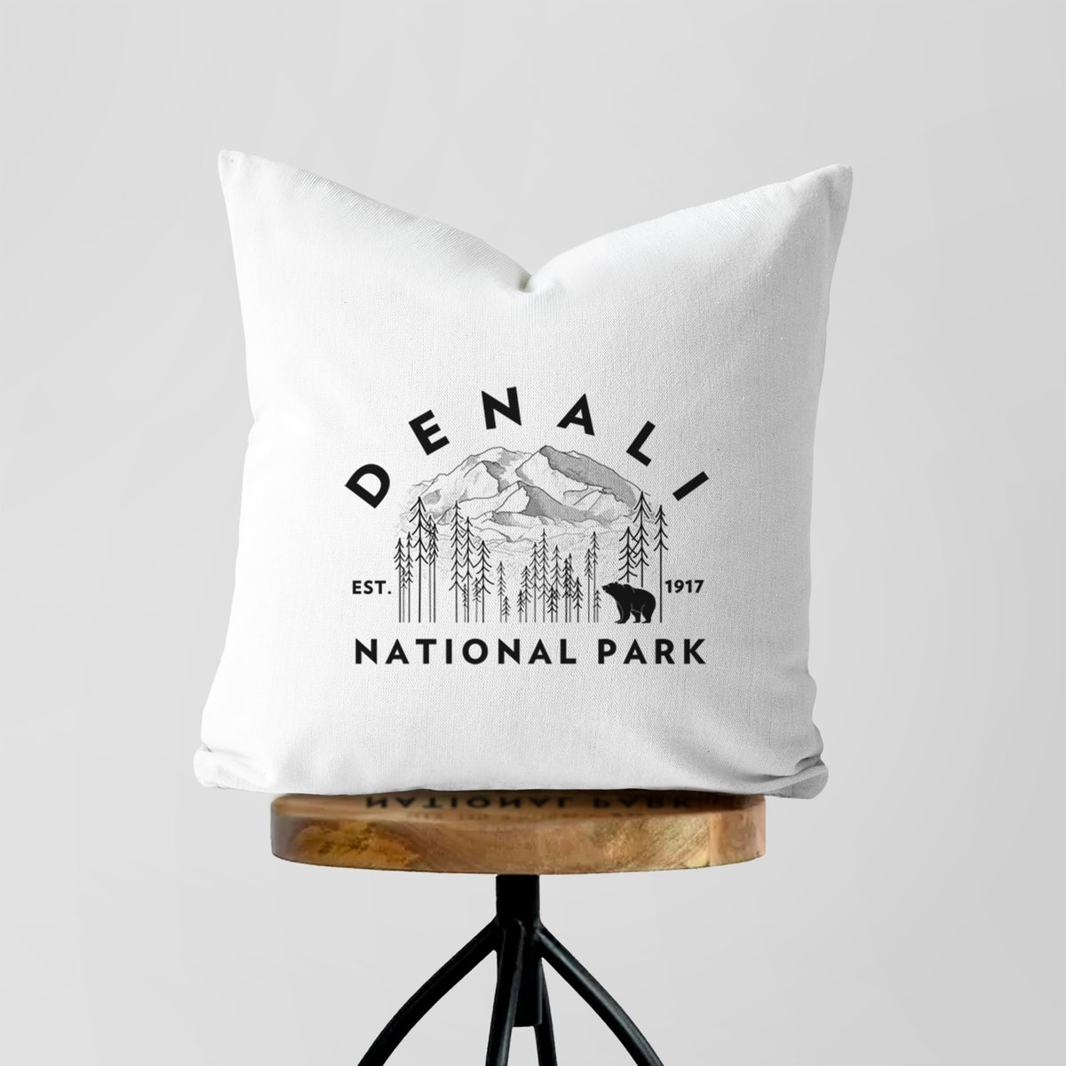 White pillow cover printed with a round badge of mountains, pine trees and a bear, with the words Denali National Park, Est. 1917, on a wooden stool.