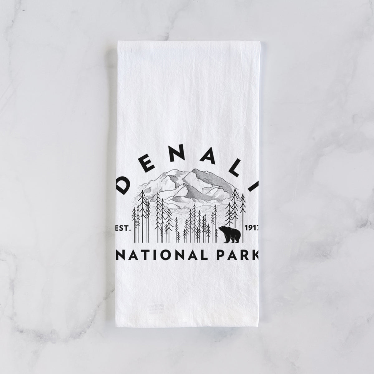 White cotton tea towel laid flat on marble, printed with a round badge of mountains, pine trees and a bear, with the words Denali National Park, Est. 1917.