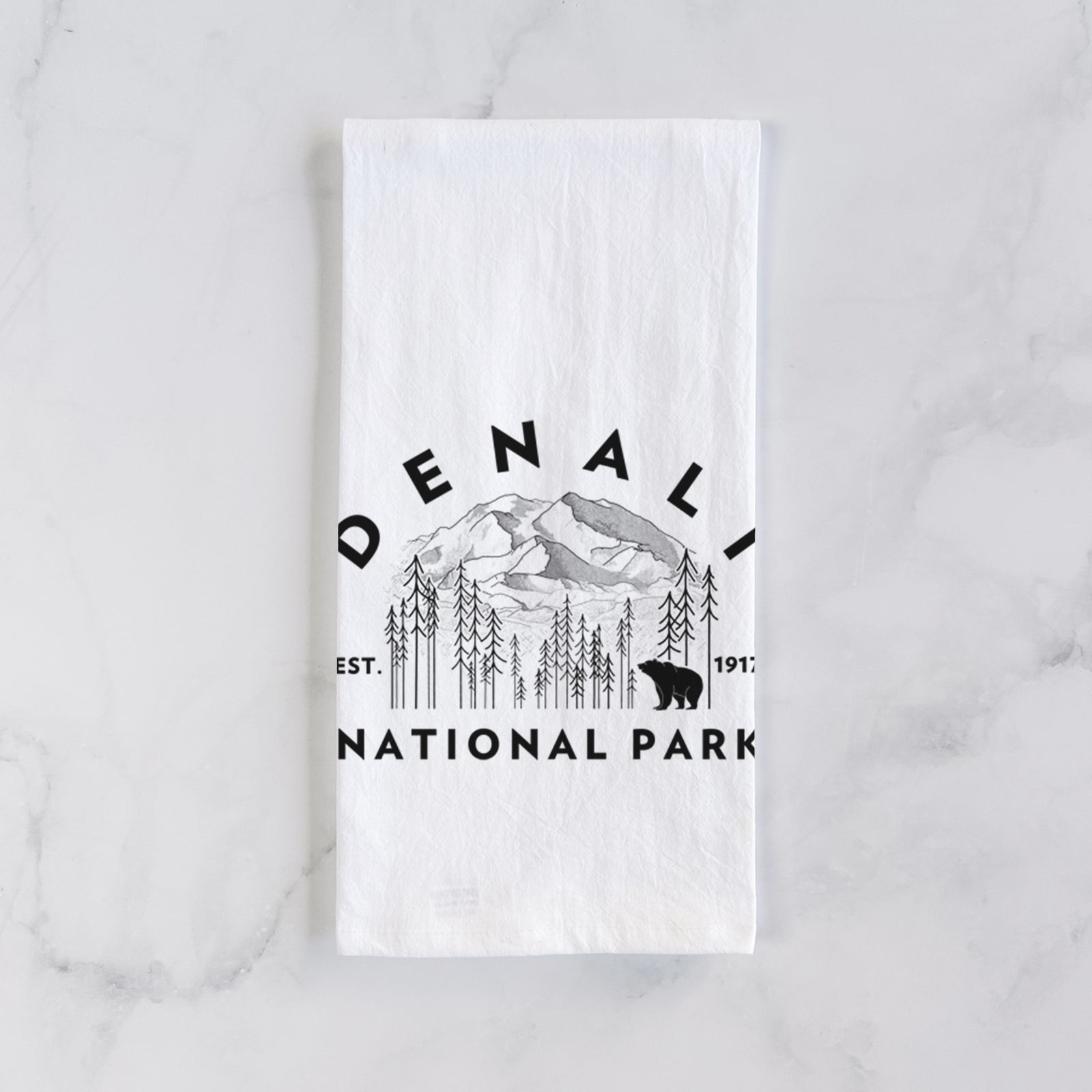 White cotton tea towel printed with a round badge of mountains, pine trees and a bear, with the words Denali National Park, Est. 1917, on a marble counter beside a round wooden cutting board and greenery.
