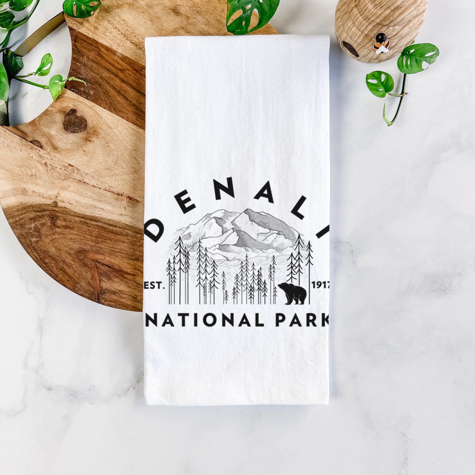 White cotton tea towel printed with a round badge of mountains, pine trees and a bear, with the words Denali National Park, Est. 1917, on a marble counter beside a round wooden cutting board and greenery.