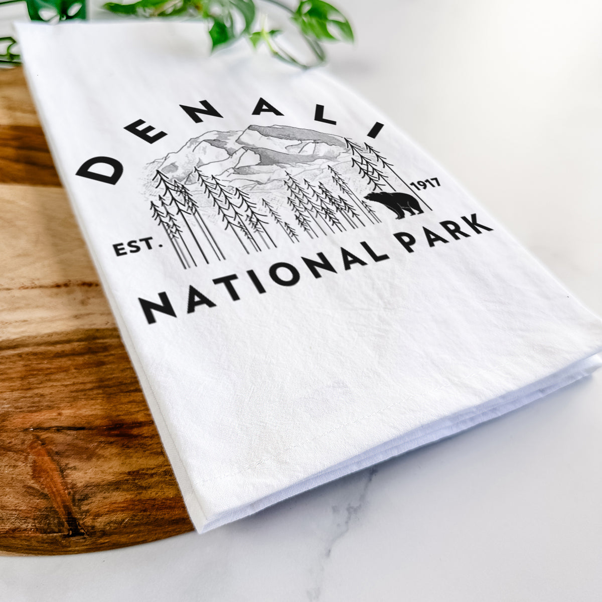 Close-up of a folded white tea towel on a wooden board, showing a round badge of mountains, pine trees and a bear, with the words Denali National Park, Est. 1917.