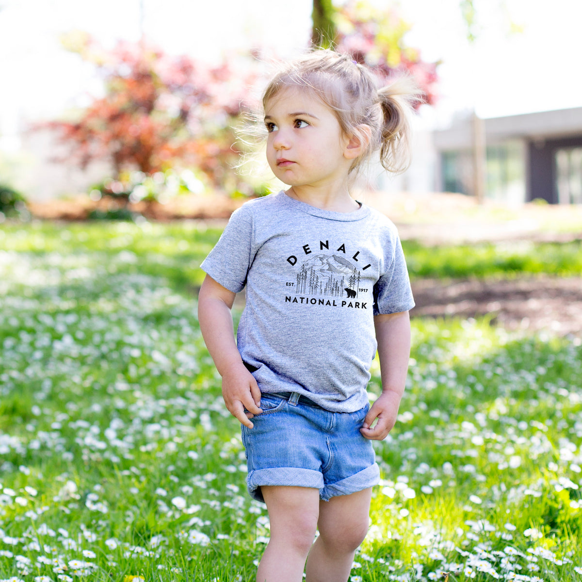 Toddler girl walking outdoors wearing a t-shirt printed with a round badge of mountains, pine trees and a bear, with the words Denali National Park, Est. 1917.