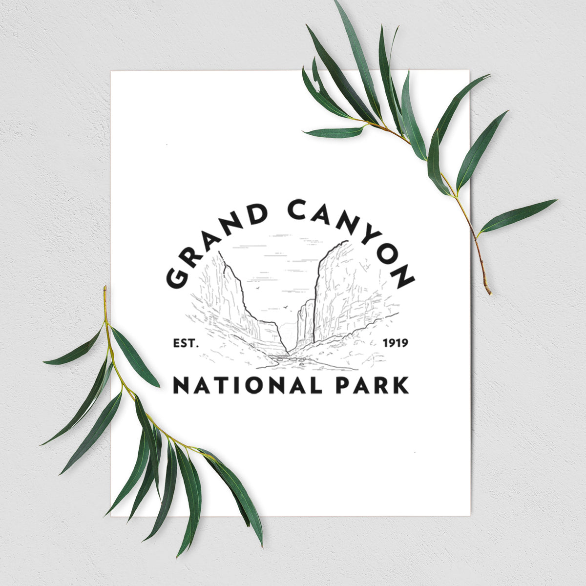 Art print of a round badge of canyon cliffs, with the words Grand Canyon National Park, Est. 1919, shown with eucalyptus leaves.