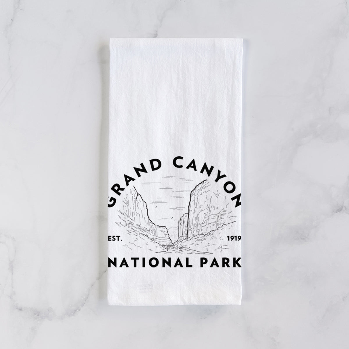 White cotton tea towel laid flat on marble, printed with a round badge of canyon cliffs, with the words Grand Canyon National Park, Est. 1919.