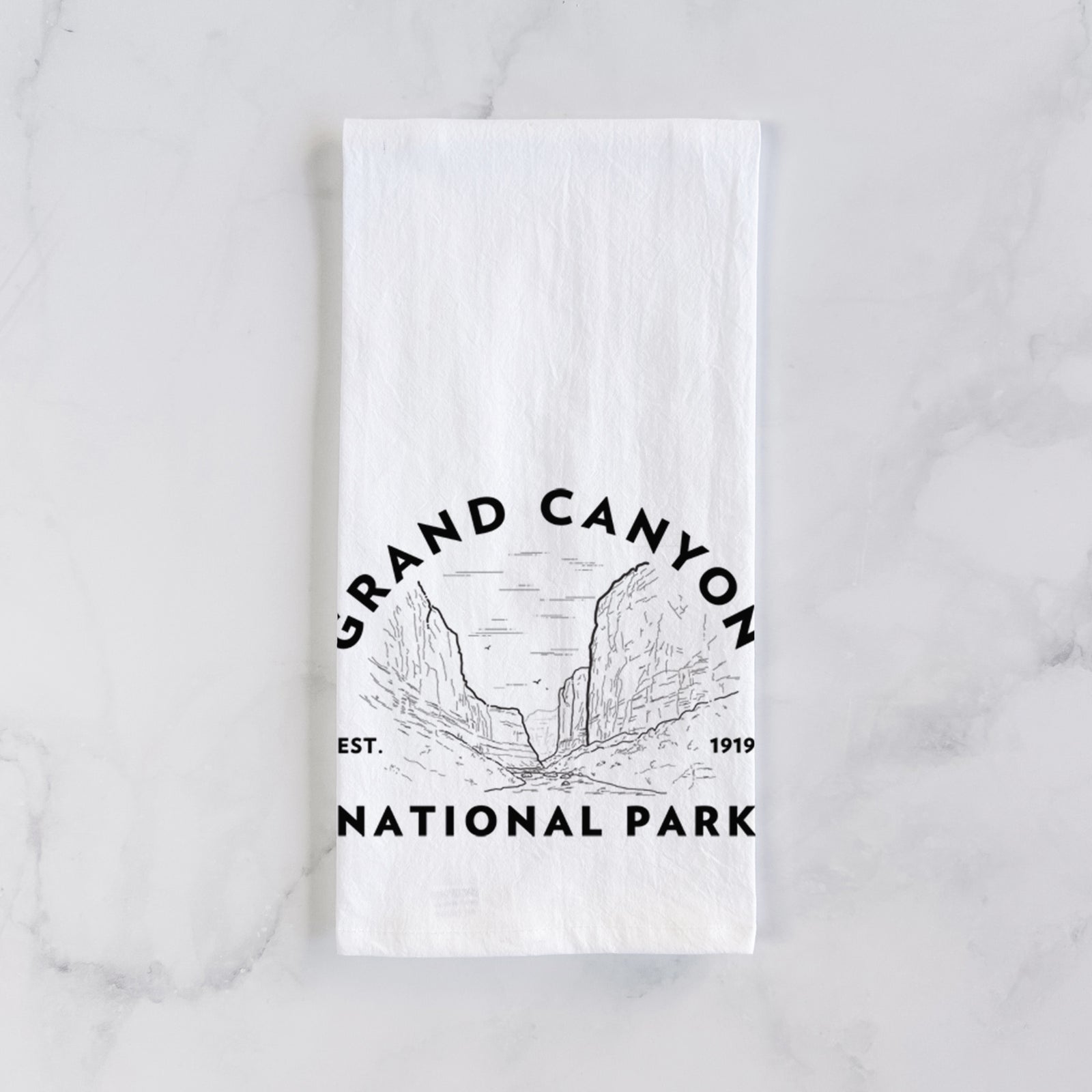 White cotton tea towel printed with a round badge of canyon cliffs, with the words Grand Canyon National Park, Est. 1919, on a marble counter beside a round wooden cutting board and greenery.