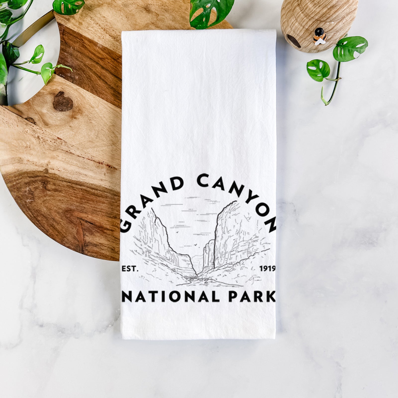 White cotton tea towel printed with a round badge of canyon cliffs, with the words Grand Canyon National Park, Est. 1919, on a marble counter beside a round wooden cutting board and greenery.