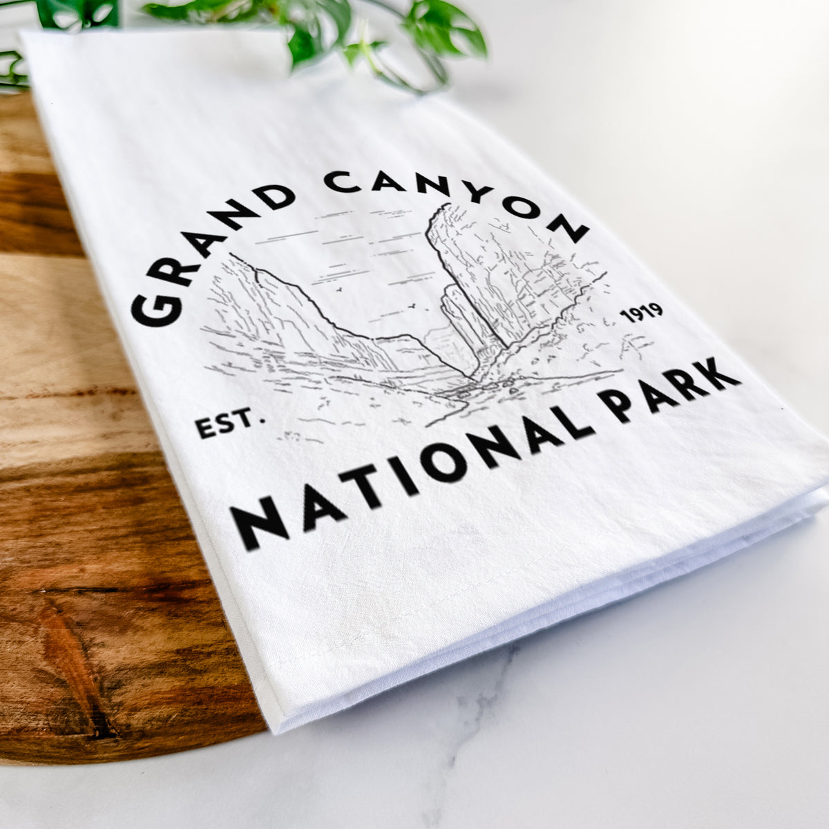 Close-up of a folded white tea towel on a wooden board, showing a round badge of canyon cliffs, with the words Grand Canyon National Park, Est. 1919.