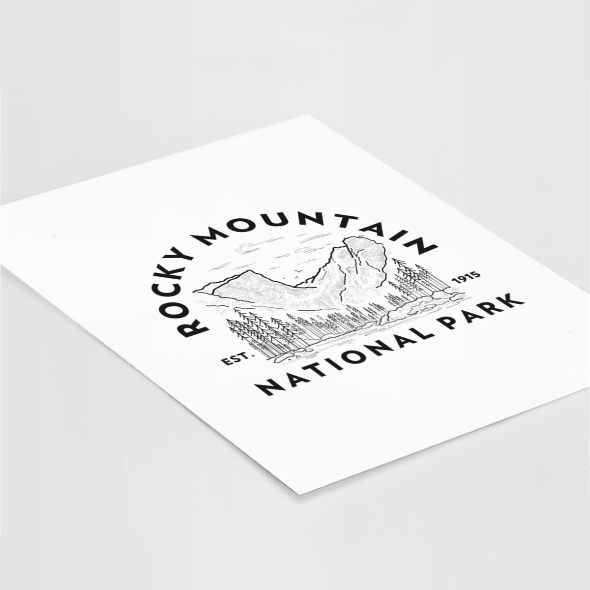 Art print of a round badge of mountains and pine trees, with the words Rocky Mountain National Park, Est. 1915, lying at an angle on a white surface.