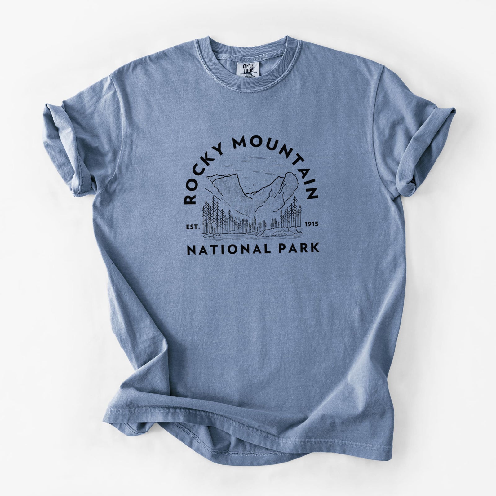Blue jean Comfort Colors t-shirt laid flat, printed with a round badge of mountains and pine trees, with the words Rocky Mountain National Park, Est. 1915.