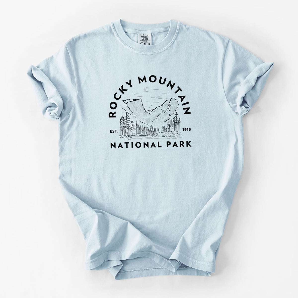 Chambray blue Comfort Colors t-shirt laid flat, printed with a round badge of mountains and pine trees, with the words Rocky Mountain National Park, Est. 1915.