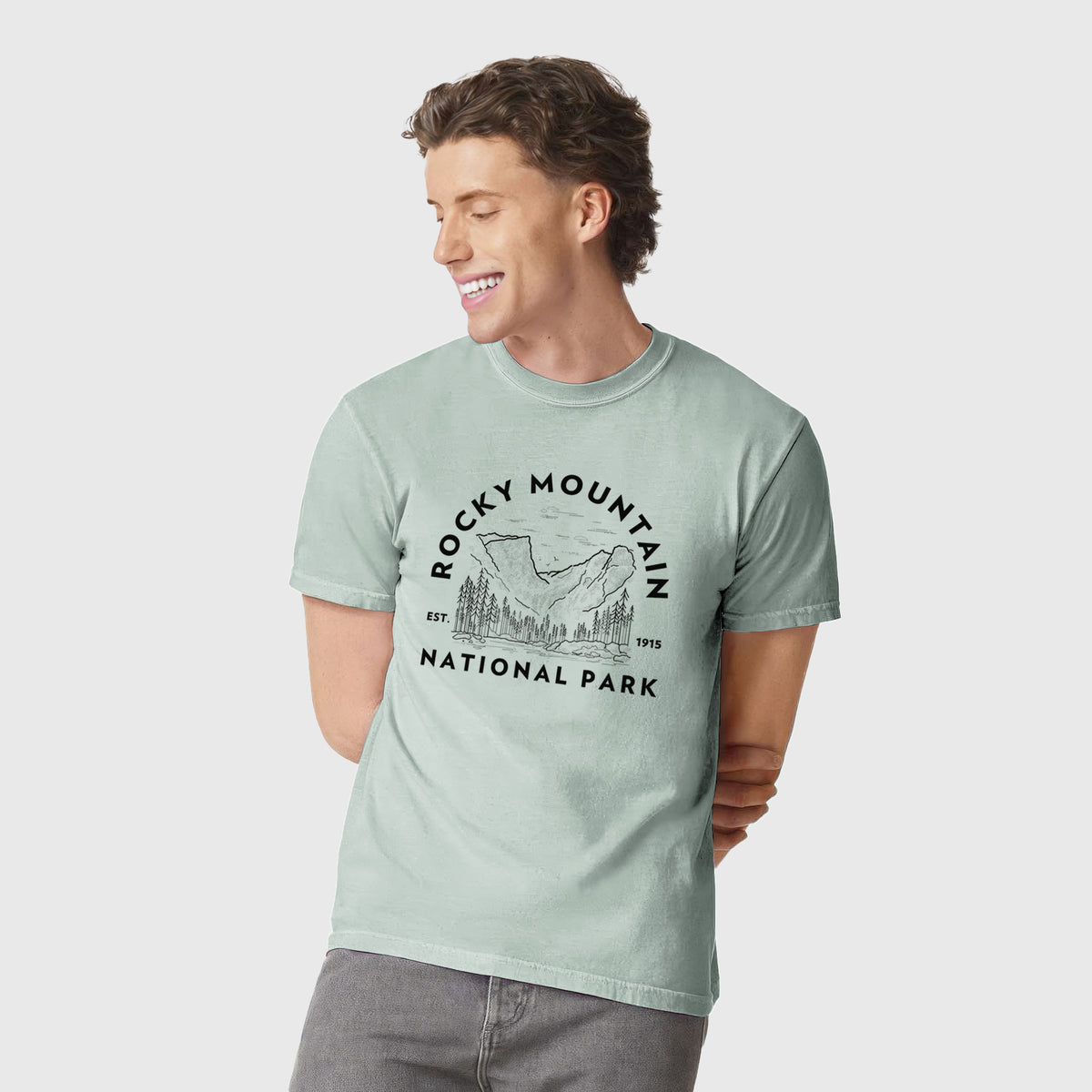 Man wearing a Comfort Colors t-shirt printed with a round badge of mountains and pine trees, with the words Rocky Mountain National Park, Est. 1915.