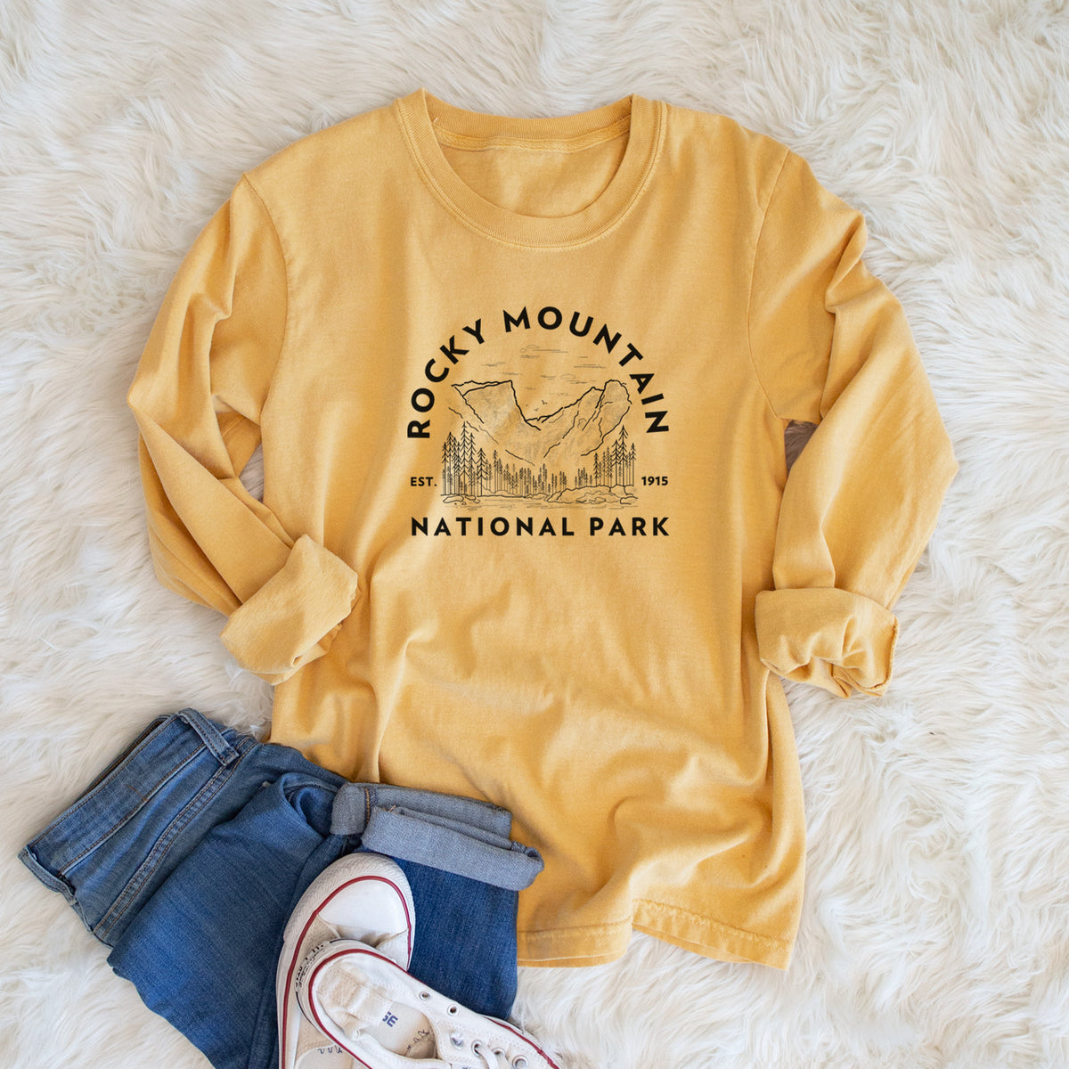 Golden wheat yellow Comfort Colors long sleeve shirt printed with a round badge of mountains and pine trees, with the words Rocky Mountain National Park, Est. 1915, shown in a lifestyle photo.