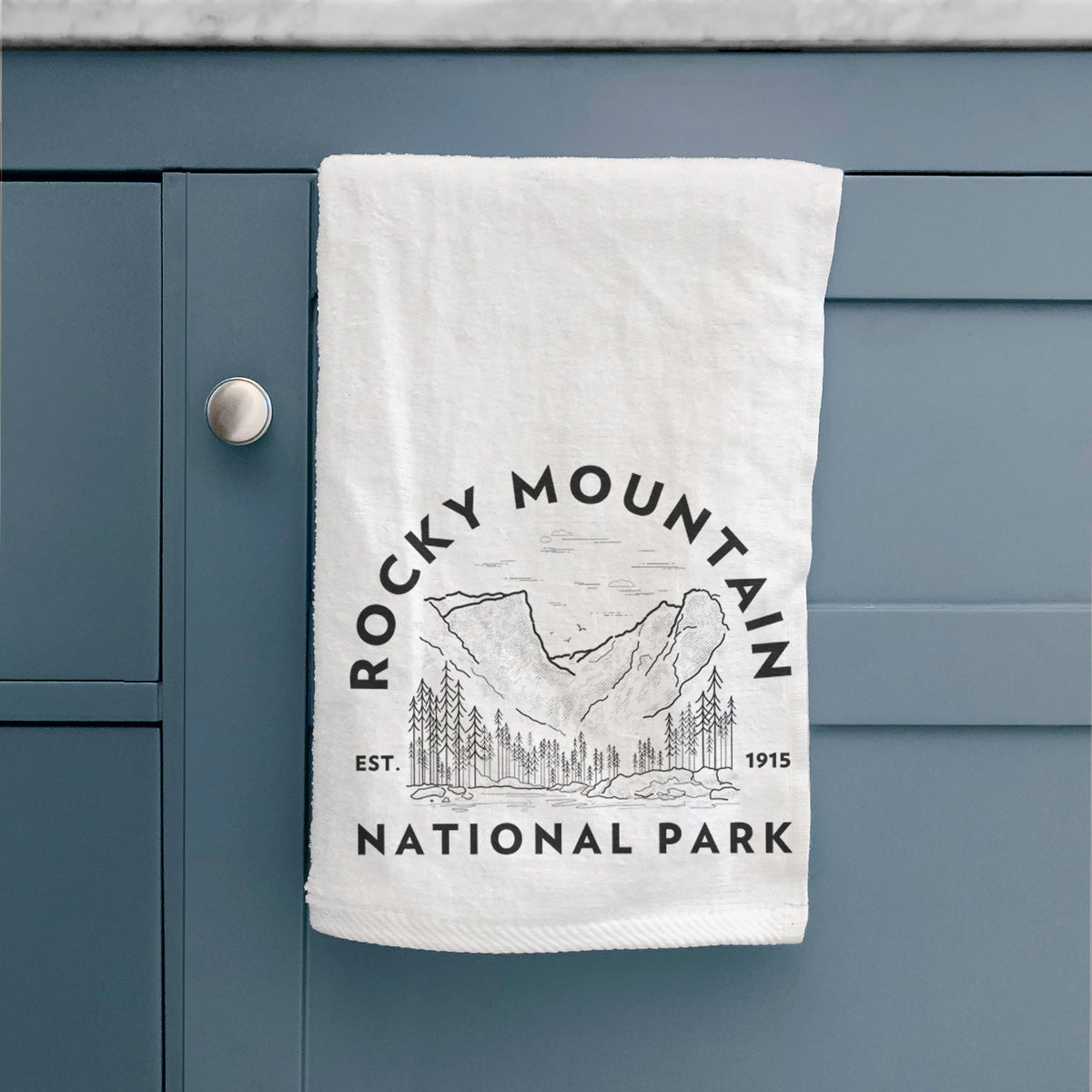 White hand towel printed with a round badge of mountains and pine trees, with the words Rocky Mountain National Park, Est. 1915, hanging on a cabinet door.