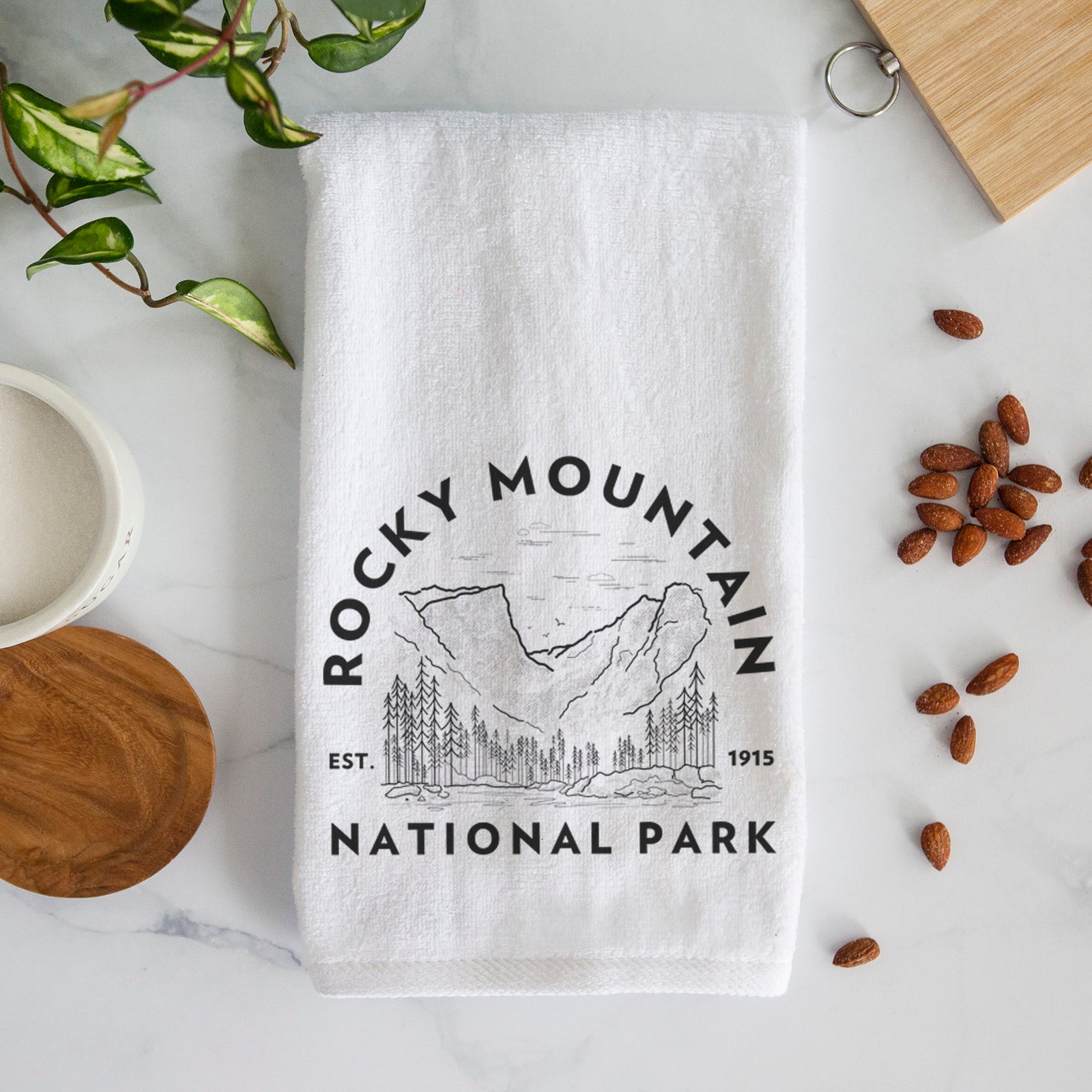 White hand towel printed with a round badge of mountains and pine trees, with the words Rocky Mountain National Park, Est. 1915.