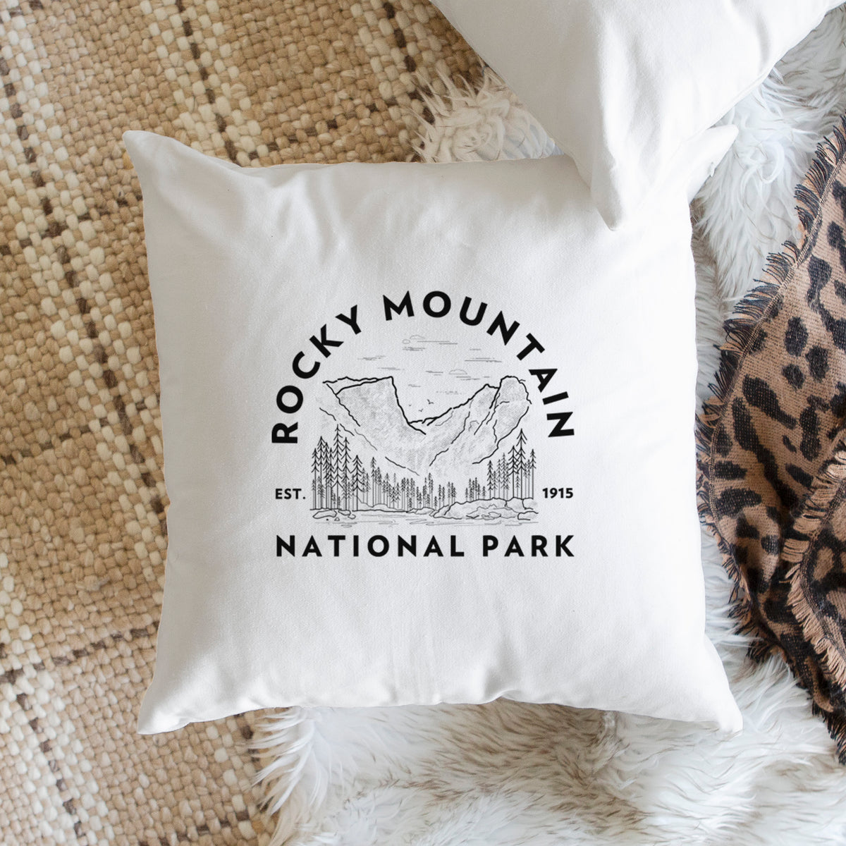 White pillow cover printed with a round badge of mountains and pine trees, with the words Rocky Mountain National Park, Est. 1915, on a woven chair.