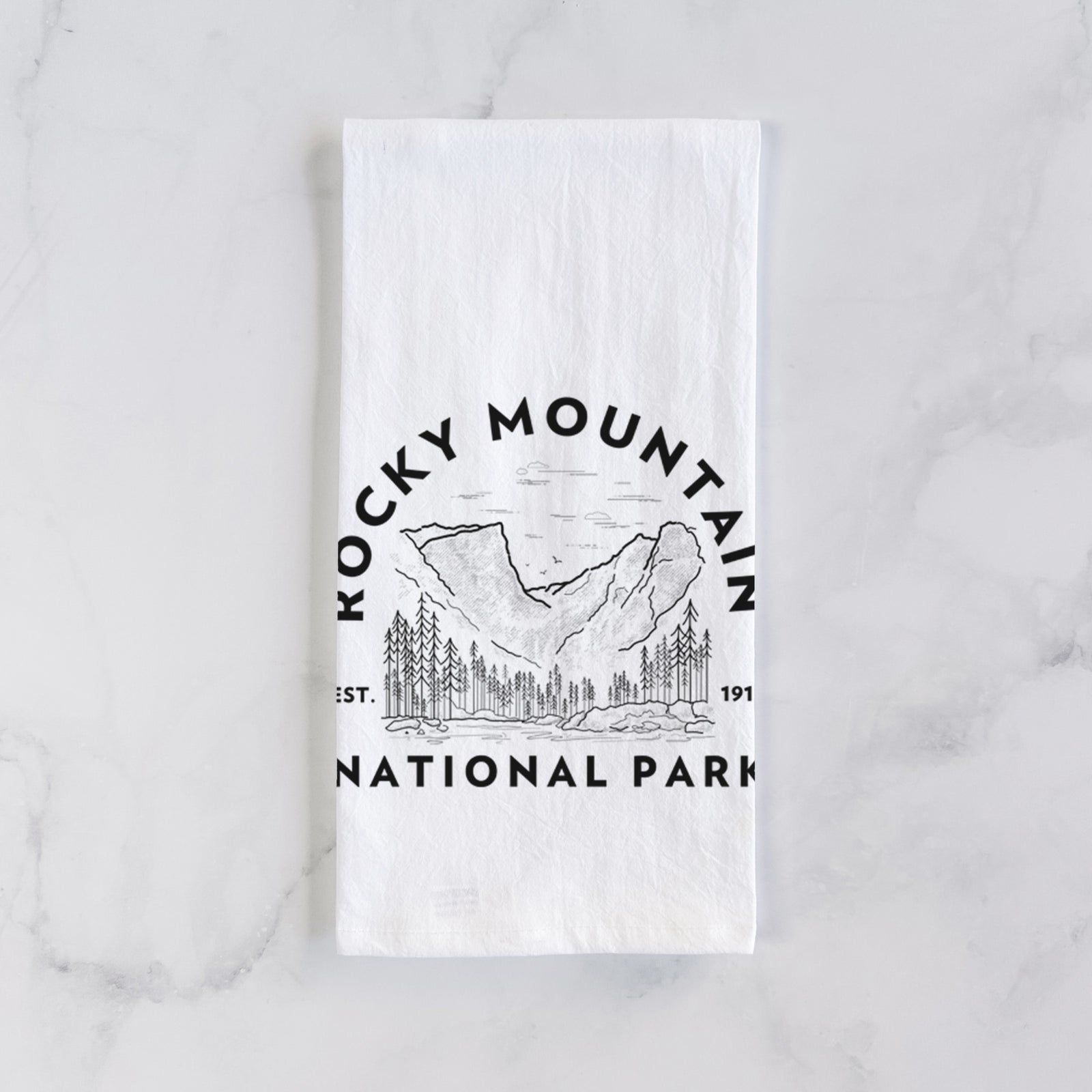 White cotton tea towel printed with a round badge of mountains and pine trees, with the words Rocky Mountain National Park, Est. 1915, on a marble counter beside a round wooden cutting board and greenery.