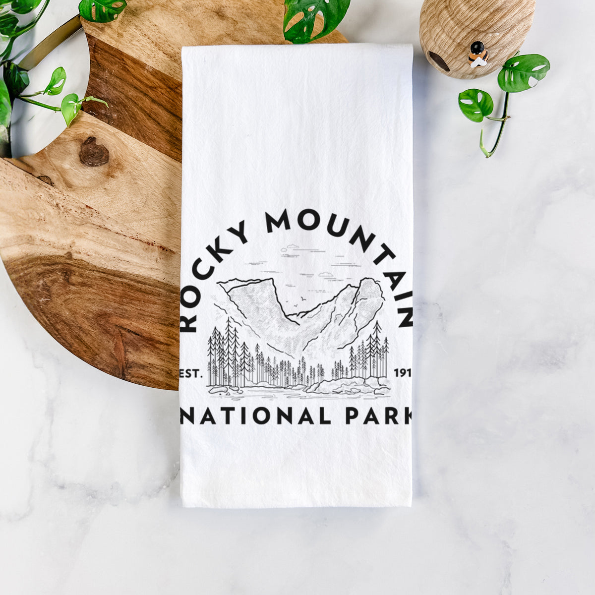White cotton tea towel printed with a round badge of mountains and pine trees, with the words Rocky Mountain National Park, Est. 1915, on a marble counter beside a round wooden cutting board and greenery.