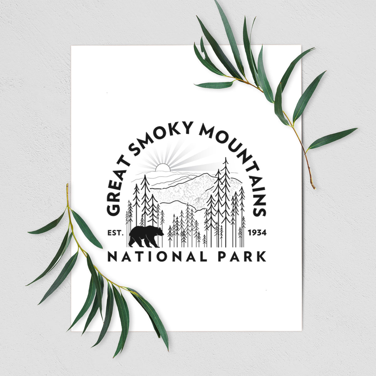 Art print of a round badge of mountains, pine trees and a black bear, with the words Great Smoky Mountains National Park, Est. 1934, shown with eucalyptus leaves.