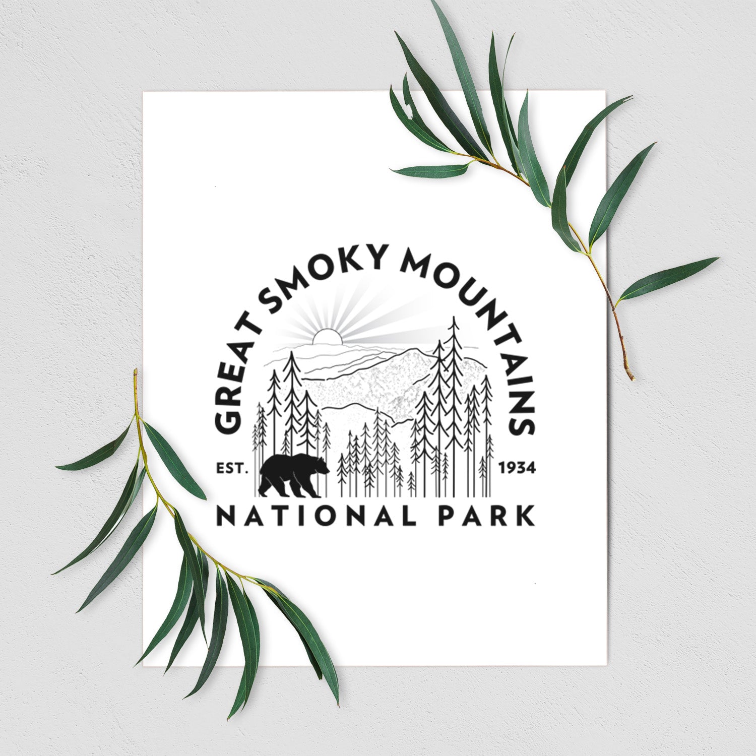 Art print of a round badge of mountains, pine trees and a black bear, with the words Great Smoky Mountains National Park, Est. 1934, shown with eucalyptus leaves.