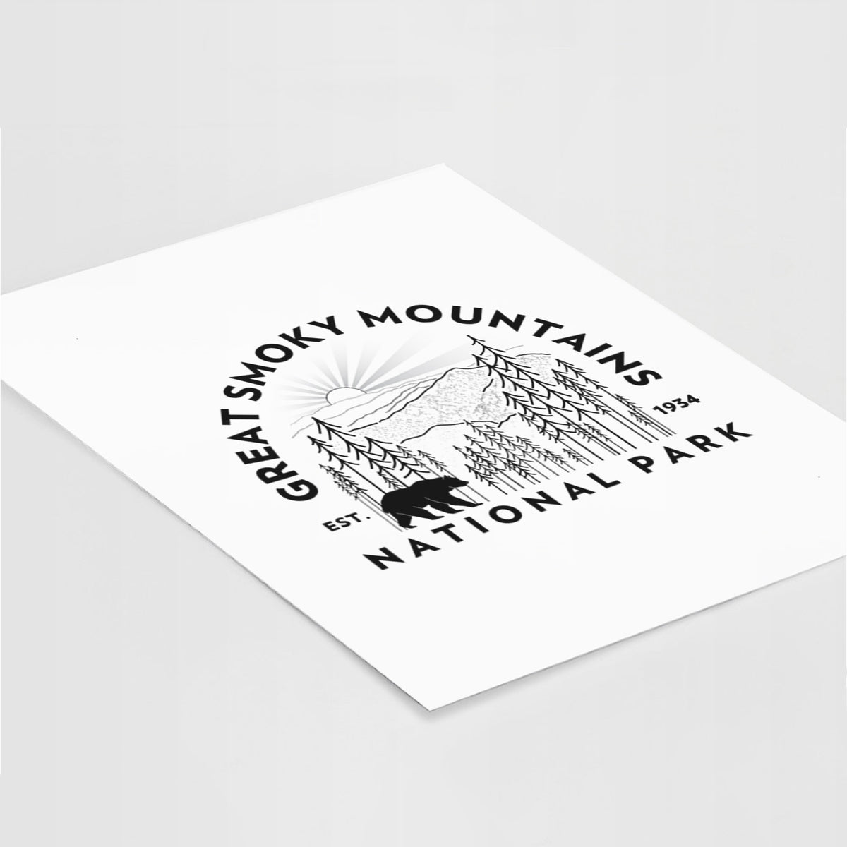 Art print of a round badge of mountains, pine trees and a black bear, with the words Great Smoky Mountains National Park, Est. 1934, lying at an angle on a white surface.
