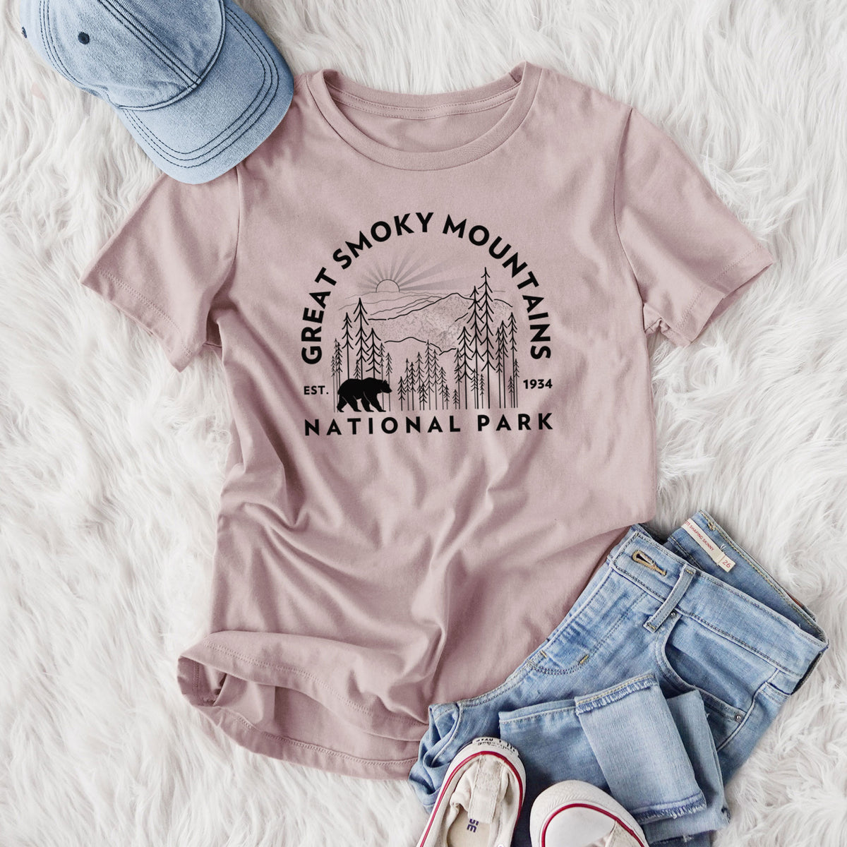 Women's relaxed fit cotton t-shirt printed with a round badge of mountains, pine trees and a black bear, with the words Great Smoky Mountains National Park, Est. 1934, styled flat on a white faux fur rug with jeans.