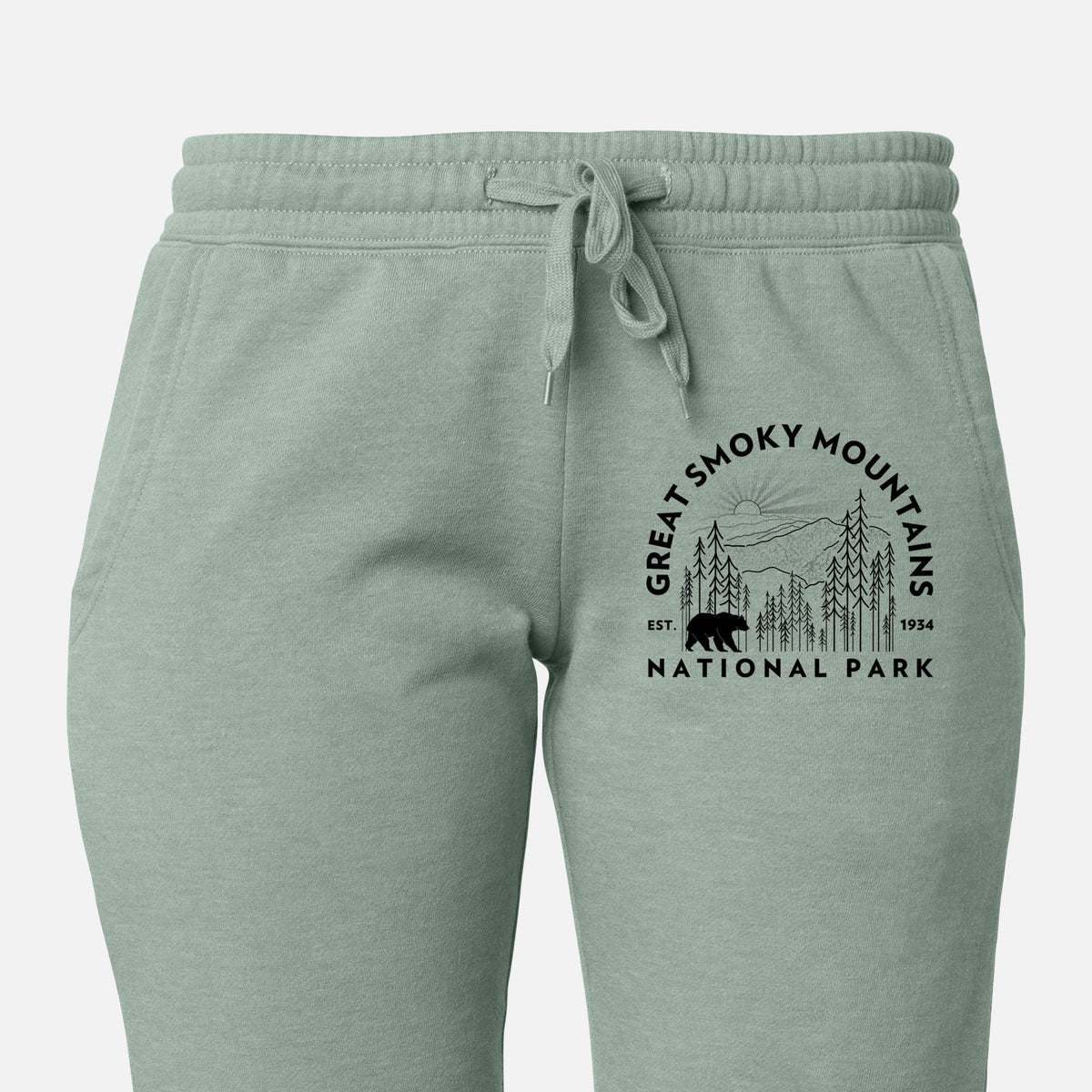 Front of sage green Cali Wave sweatpants, printed with a round badge of mountains, pine trees and a black bear, with the words Great Smoky Mountains National Park, Est. 1934 on the leg.