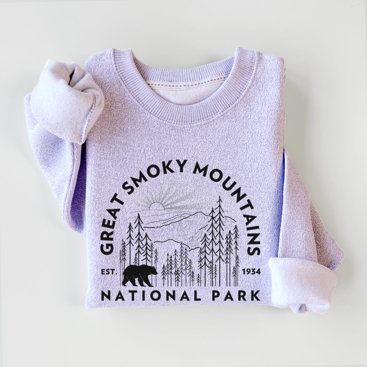 Ice purple knit sweatshirt printed with a round badge of mountains, pine trees and a black bear, with the words Great Smoky Mountains National Park, Est. 1934, styled flat.