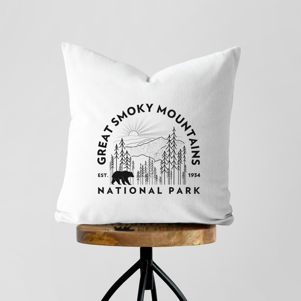White pillow cover printed with a round badge of mountains, pine trees and a black bear, with the words Great Smoky Mountains National Park, Est. 1934, on a wooden stool.
