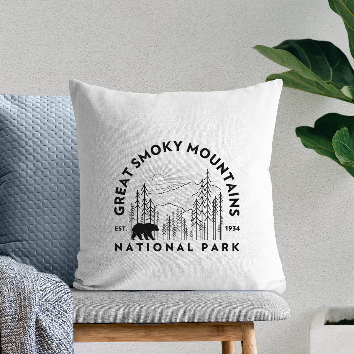 White pillow cover printed with a round badge of mountains, pine trees and a black bear, with the words Great Smoky Mountains National Park, Est. 1934, on a bench beside plants.
