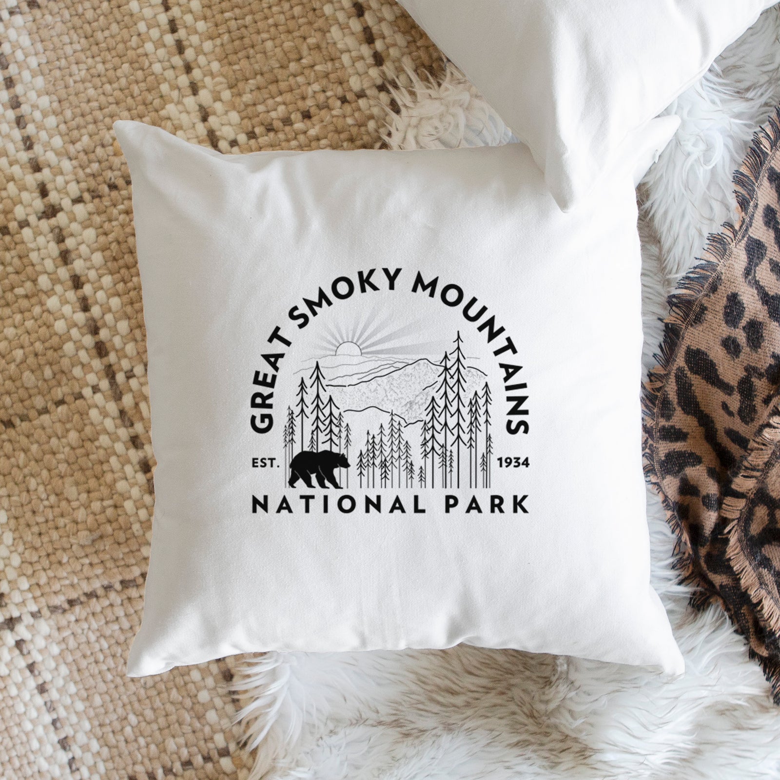 White pillow cover printed with a round badge of mountains, pine trees and a black bear, with the words Great Smoky Mountains National Park, Est. 1934, on a bench beside plants.