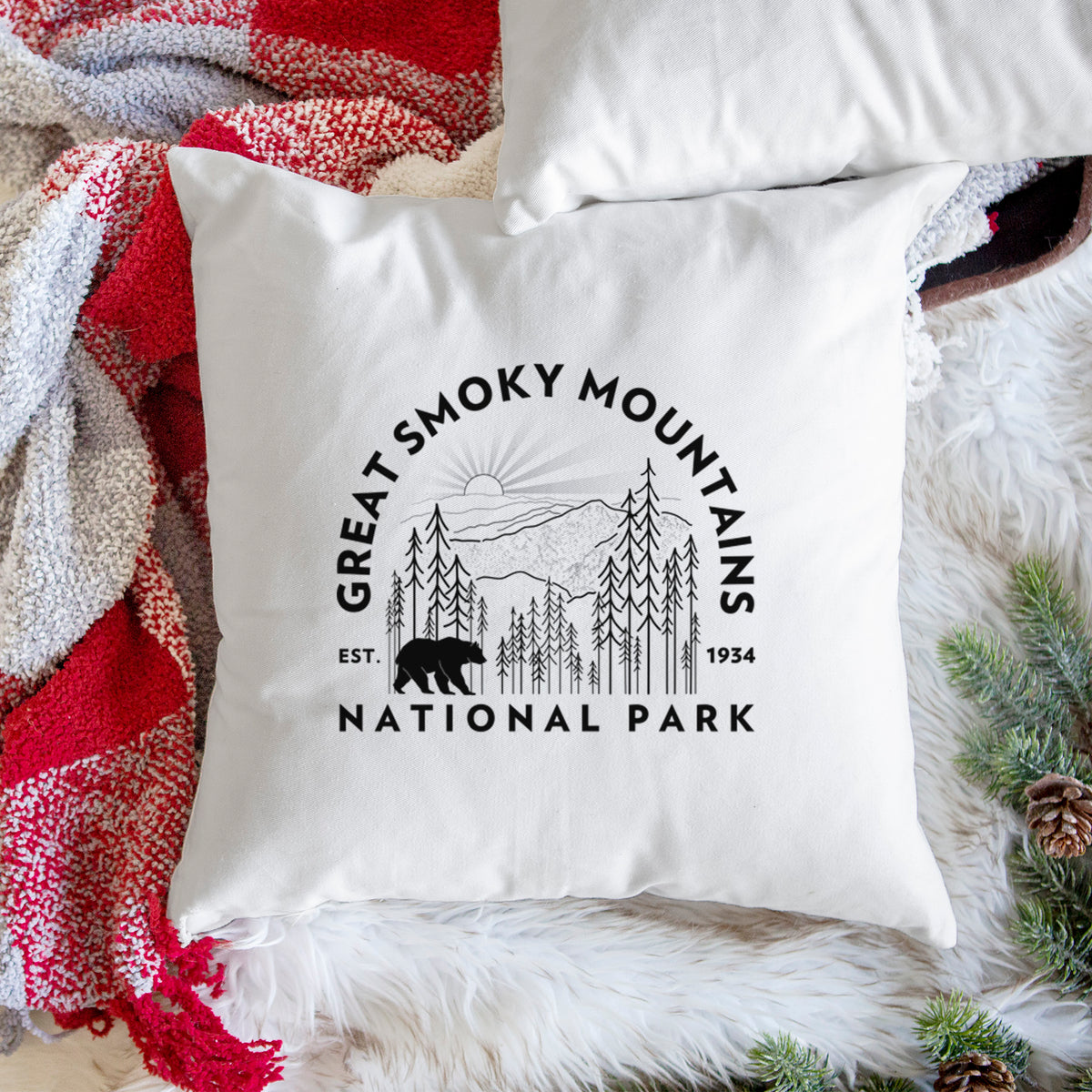 White pillow cover printed with a round badge of mountains, pine trees and a black bear, with the words Great Smoky Mountains National Park, Est. 1934, on a woven chair.
