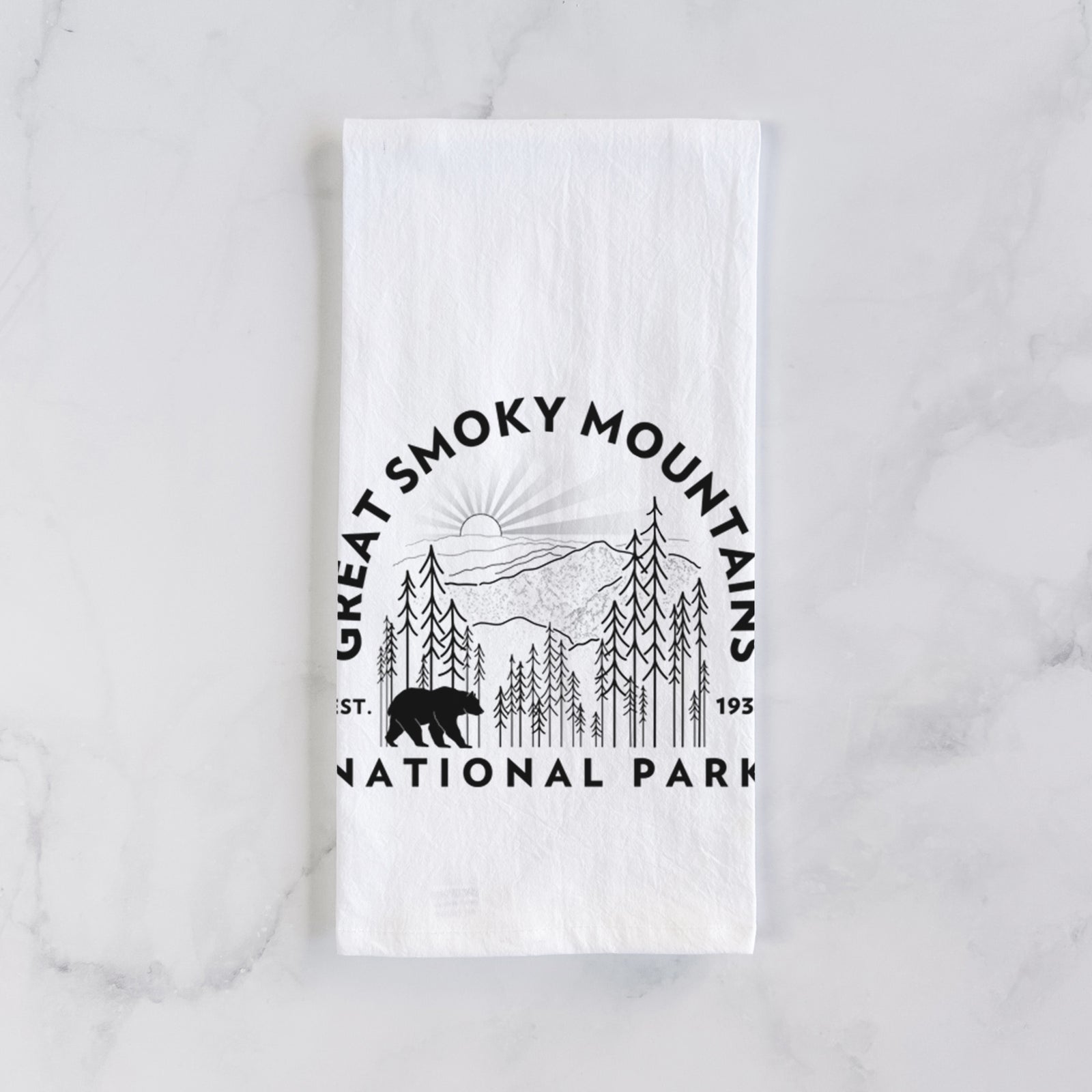 White cotton tea towel printed with a round badge of mountains, pine trees and a black bear, with the words Great Smoky Mountains National Park, Est. 1934, on a marble counter beside a round wooden cutting board and greenery.