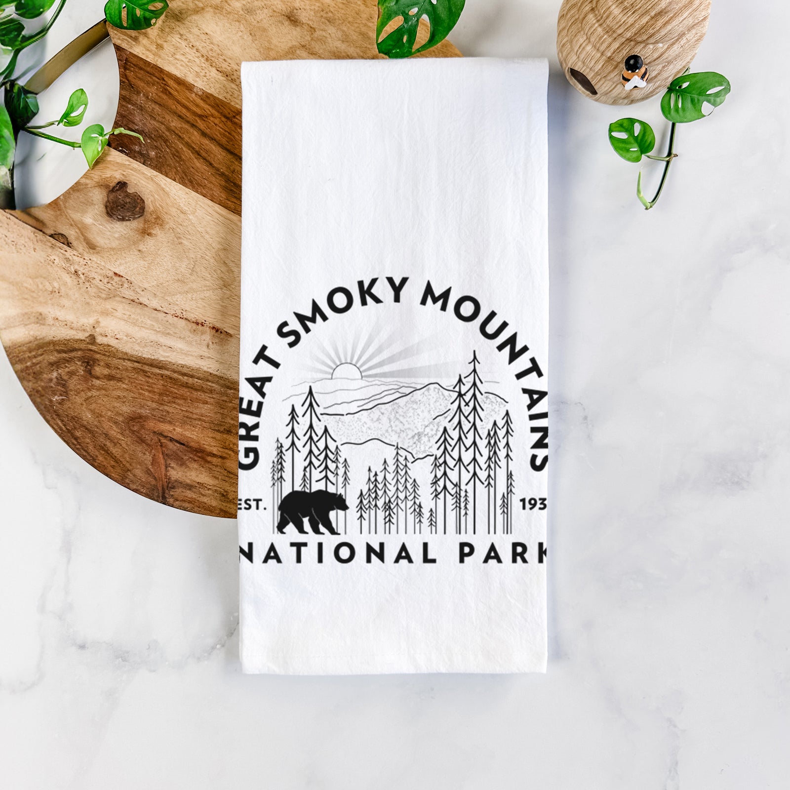 White cotton tea towel printed with a round badge of mountains, pine trees and a black bear, with the words Great Smoky Mountains National Park, Est. 1934, on a marble counter beside a round wooden cutting board and greenery.