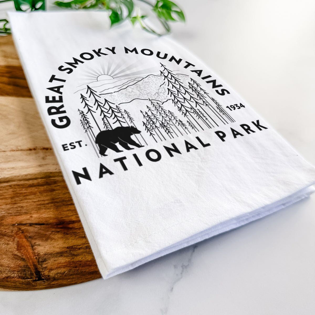 Close-up of a folded white tea towel on a wooden board, showing a round badge of mountains, pine trees and a black bear, with the words Great Smoky Mountains National Park, Est. 1934.