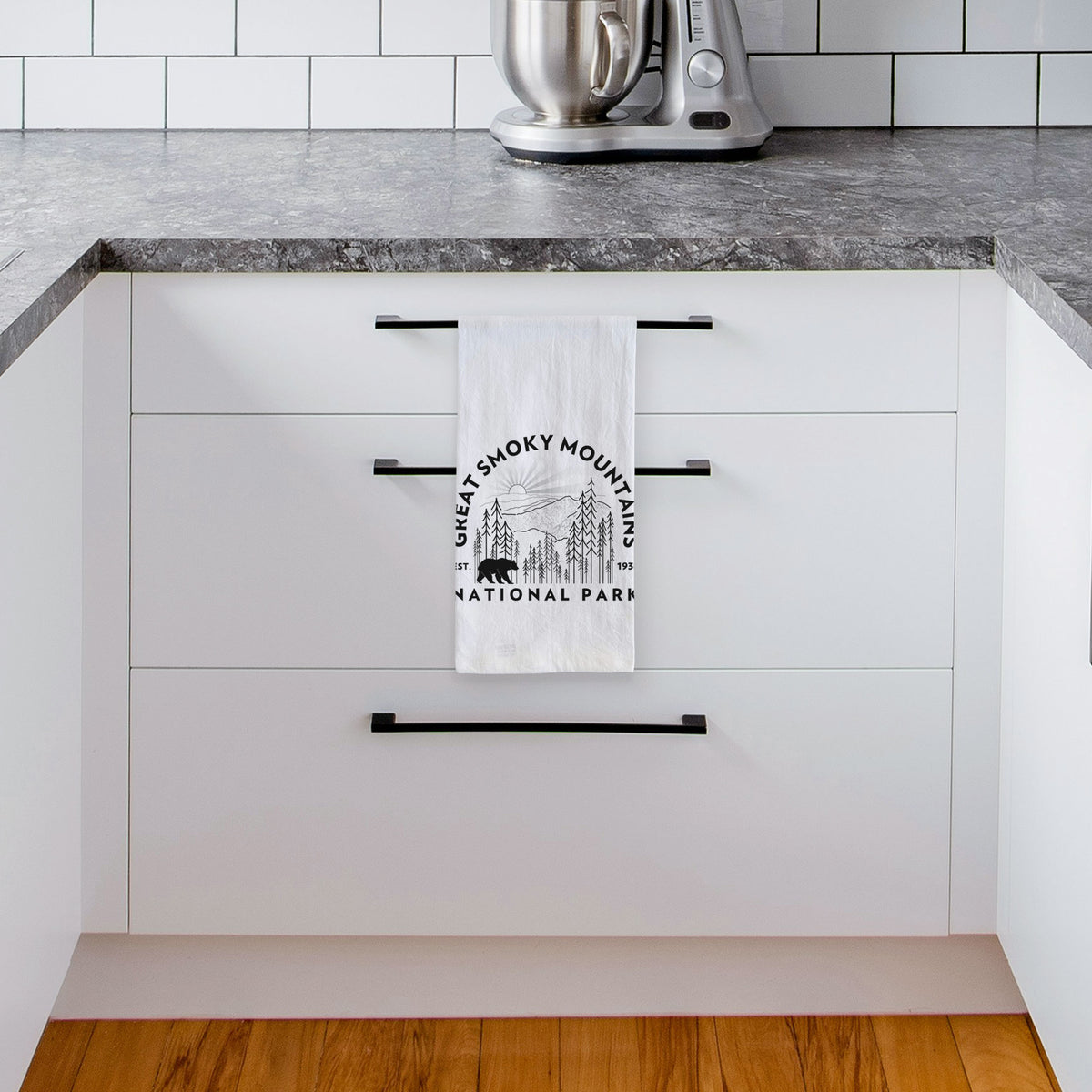 White tea towel printed with a round badge of mountains, pine trees and a black bear, with the words Great Smoky Mountains National Park, Est. 1934, hanging from a drawer handle in a bright white kitchen.
