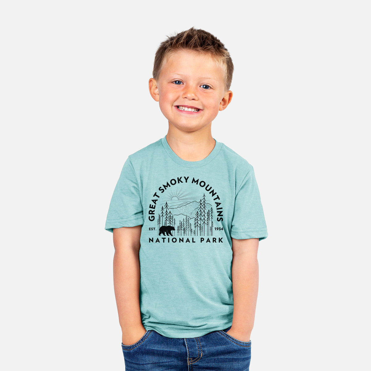 Smiling boy wearing a youth t-shirt printed with a round badge of mountains, pine trees and a black bear, with the words Great Smoky Mountains National Park, Est. 1934.