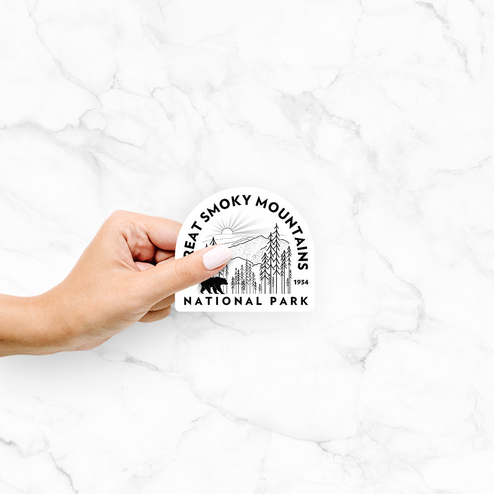 Hand holding a 4 inch vinyl decal of a round badge of mountains, pine trees and a black bear, with the words Great Smoky Mountains National Park, Est. 1934.