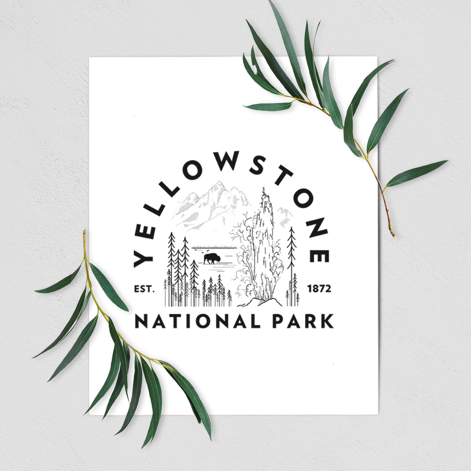 Art print of a round badge of a waterfall, pine trees and a bison, with the words Yellowstone National Park, Est. 1872, shown with eucalyptus leaves.