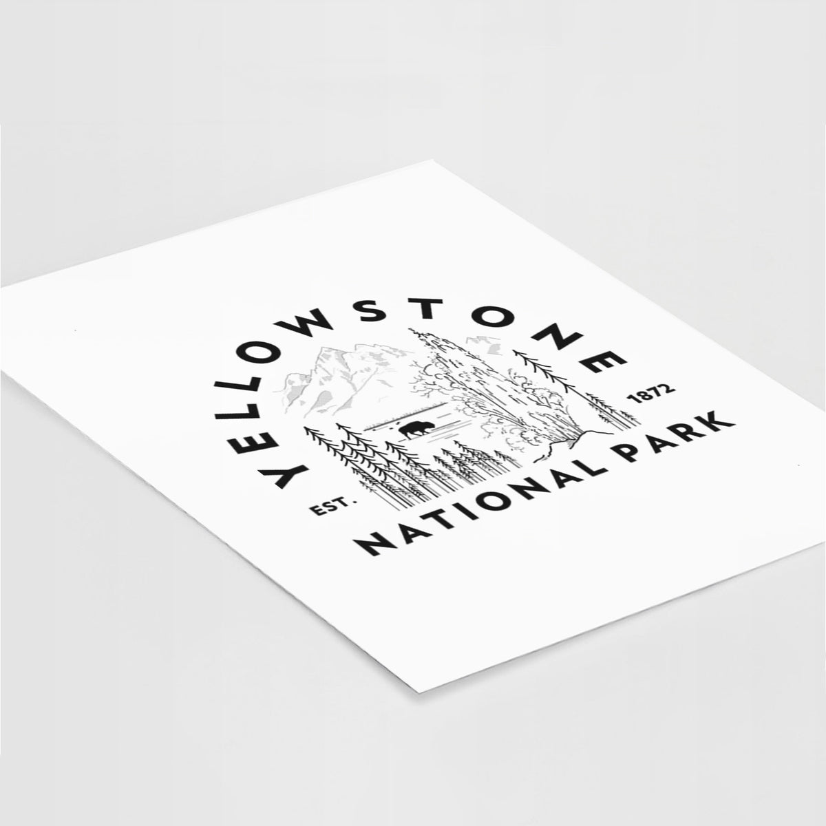 Art print of a round badge of a waterfall, pine trees and a bison, with the words Yellowstone National Park, Est. 1872, lying at an angle on a white surface.