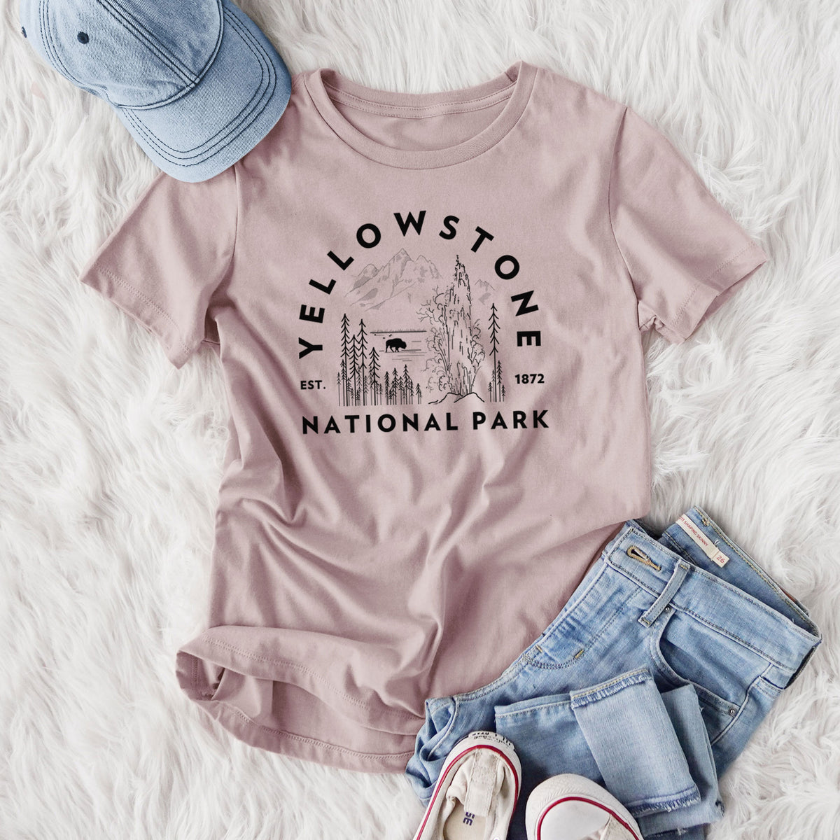 Women's relaxed fit cotton t-shirt printed with a round badge of a waterfall, pine trees and a bison, with the words Yellowstone National Park, Est. 1872, styled flat on a white faux fur rug with jeans.