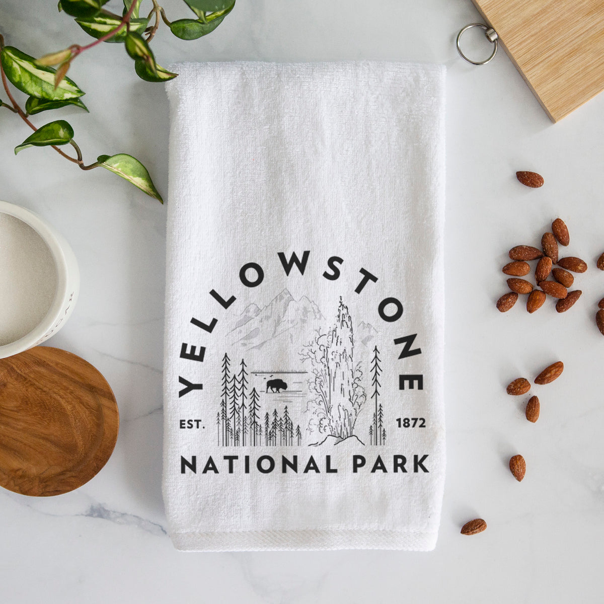 White hand towel printed with a round badge of a waterfall, pine trees and a bison, with the words Yellowstone National Park, Est. 1872.