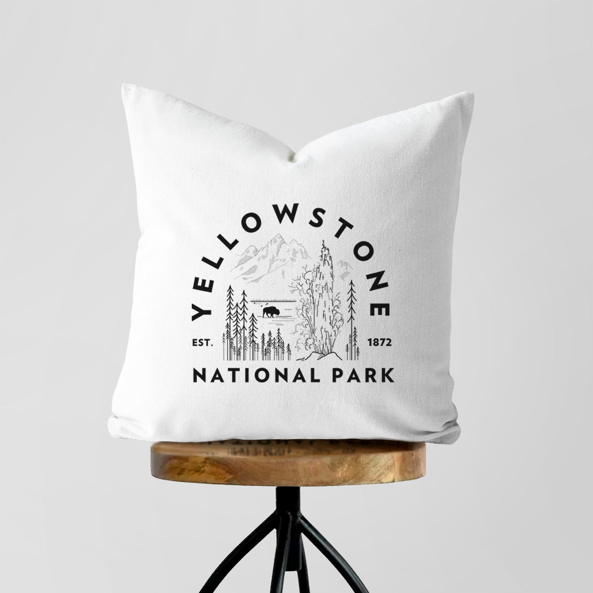 White pillow cover printed with a round badge of a waterfall, pine trees and a bison, with the words Yellowstone National Park, Est. 1872, on a wooden stool.