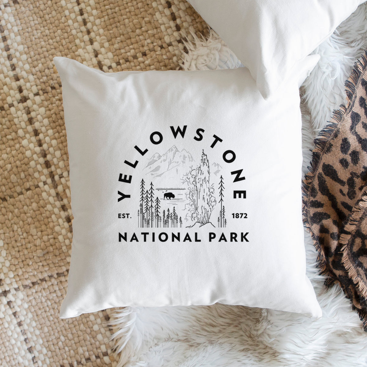 White pillow cover printed with a round badge of a waterfall, pine trees and a bison, with the words Yellowstone National Park, Est. 1872, on a woven chair.