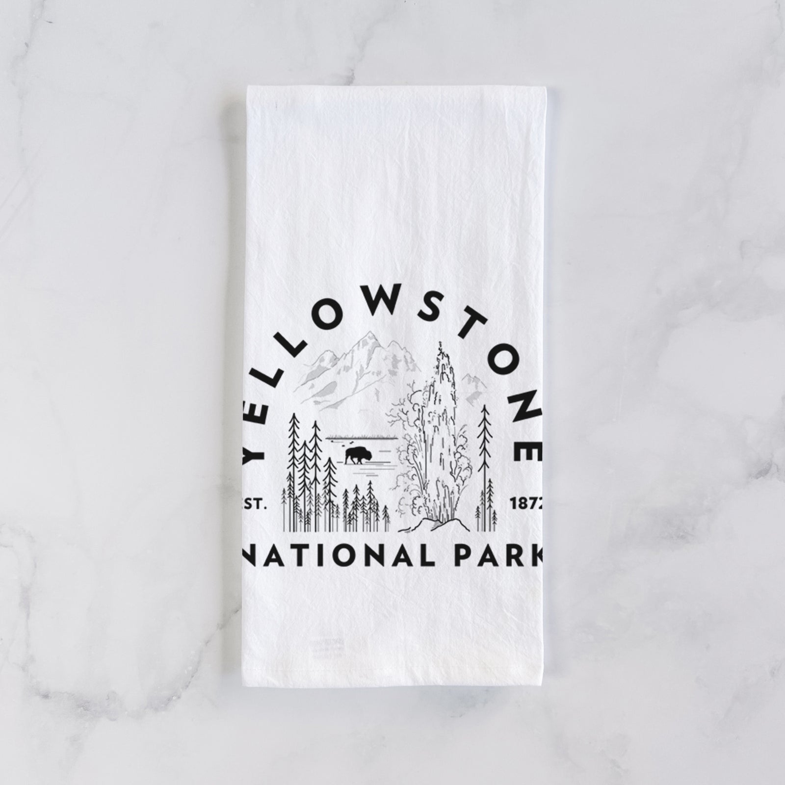 White cotton tea towel printed with a round badge of a waterfall, pine trees and a bison, with the words Yellowstone National Park, Est. 1872, on a marble counter beside a round wooden cutting board and greenery.