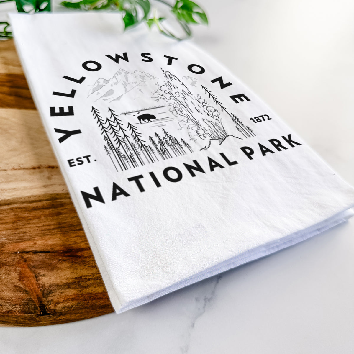 Close-up of a folded white tea towel on a wooden board, showing a round badge of a waterfall, pine trees and a bison, with the words Yellowstone National Park, Est. 1872.