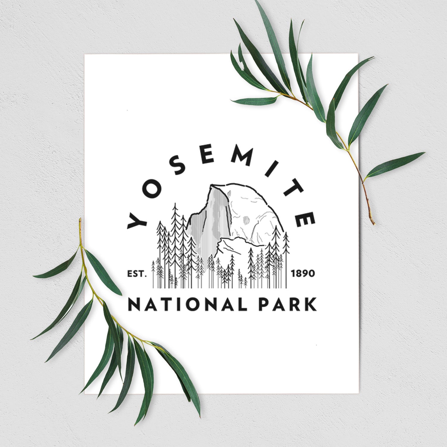 Art print of a round badge of Half Dome and pine trees, with the words Yosemite National Park, Est. 1890, shown with eucalyptus leaves.