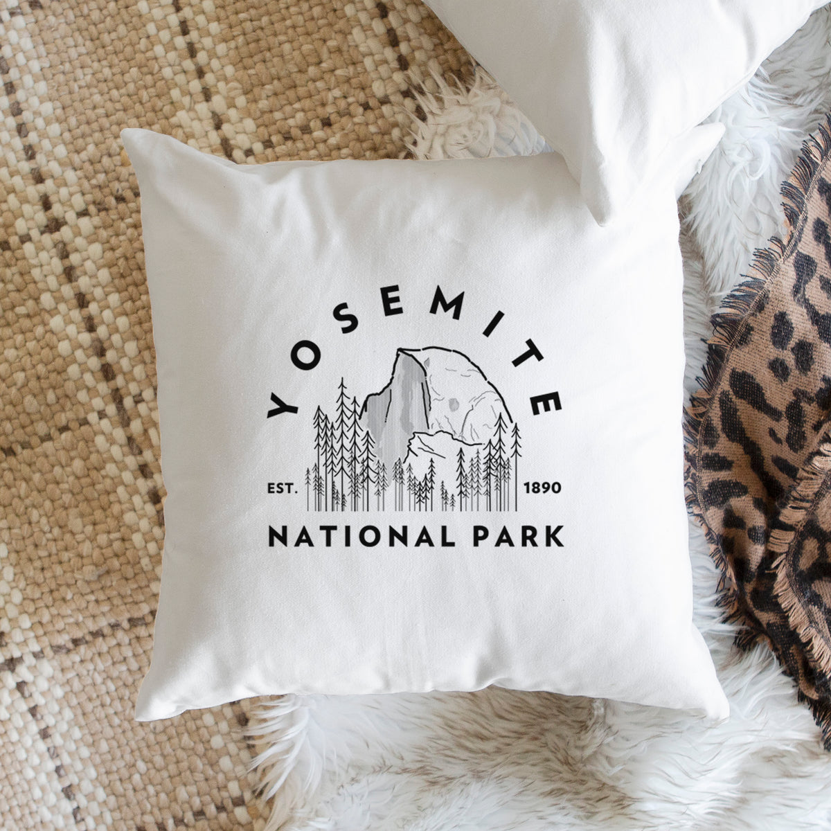 White pillow cover printed with a round badge of Half Dome and pine trees, with the words Yosemite National Park, Est. 1890, on a woven chair.