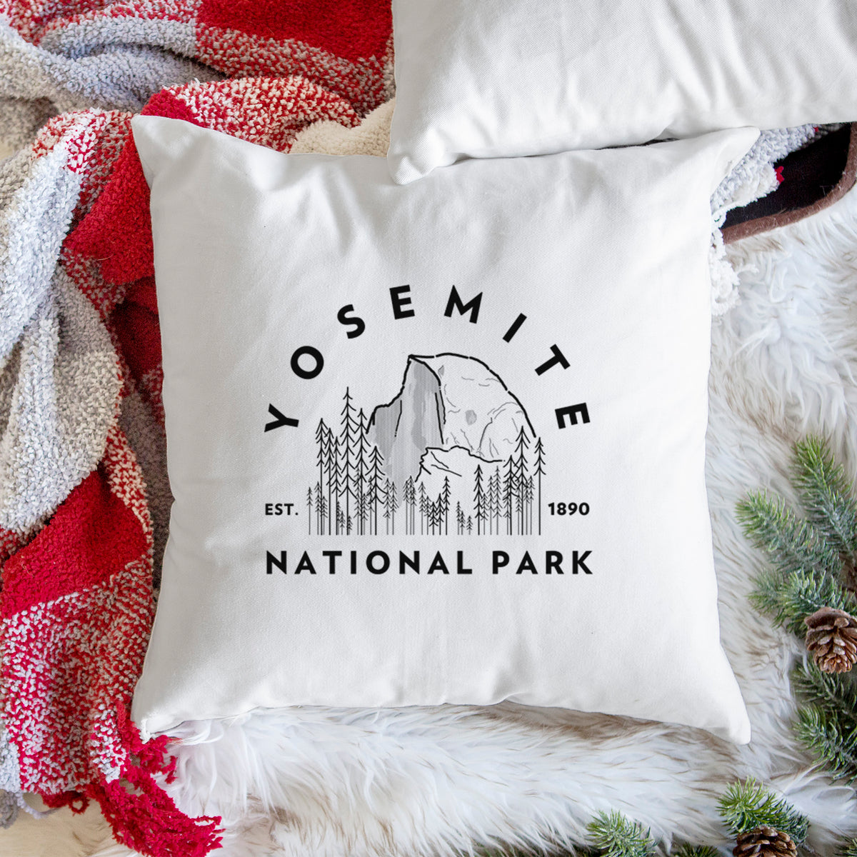 White pillow cover printed with a round badge of Half Dome and pine trees, with the words Yosemite National Park, Est. 1890, on a woven chair.